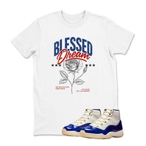May include: White t-shirt with the words "Blessed Dream" in blue and red, a rose graphic, and text at the bottom. A pair of blue, white, and black sneakers are in front of the shirt.