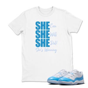 May include: White t-shirt with the text "SHE Can SHE Will SHE Did! She's Winning" in blue. A pair of light blue and white sneakers are in the foreground.