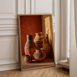 May include: A framed print depicting a selection of terracotta vases. The vases showcase diverse sizes and designs, including geometric patterns and textured surfaces. The backdrop is a warm, reddish-orange colour, and the print rests on a wooden floor.