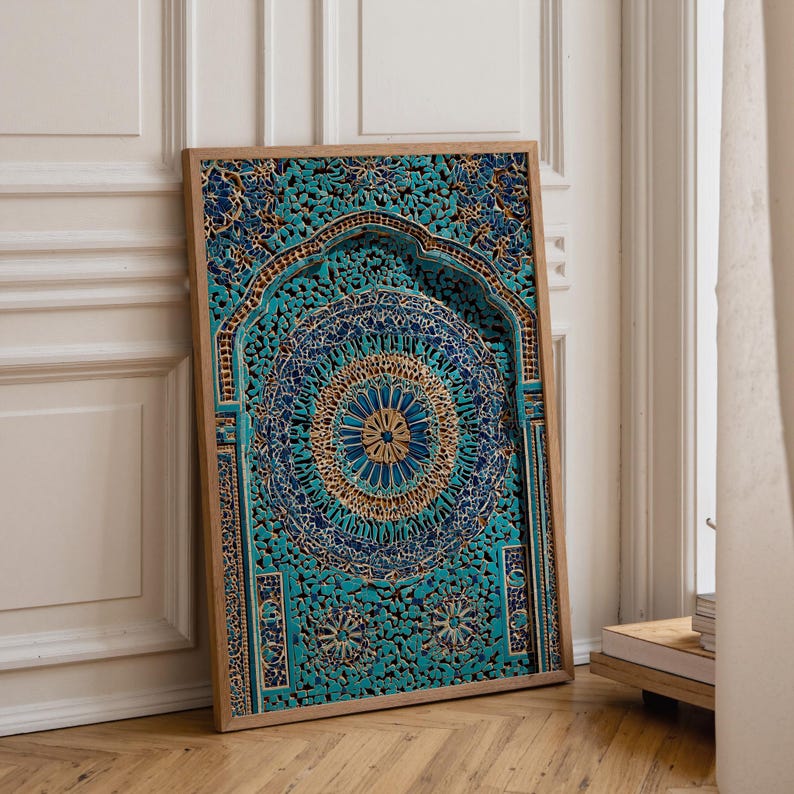 May include: A framed wooden panel with an elaborate carved design in turquoise, blue, and gold. The artwork features a mandala-style pattern with a central circular motif. The frame is a natural wood colour. The panel is leaning against a white wall and wooden floor.