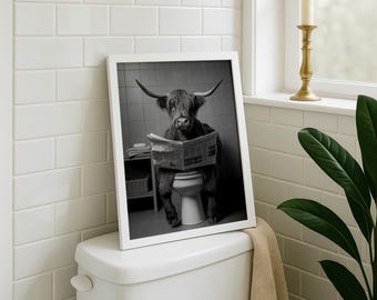 May include: A black and white framed wall art print featuring a Highland cow with long horns sitting on a toilet while reading a newspaper. The cow has shaggy fur covering its eyes and is positioned in a tiled bathroom setting with a small shelf.