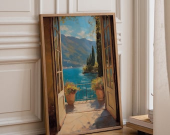 May include: A framed vertical wall art print depicting an open doorway looking out onto a balcony with terracotta pots containing flowers, overlooking a blue lake surrounded by mountains under a bright sky with tall cypress trees in the distance.