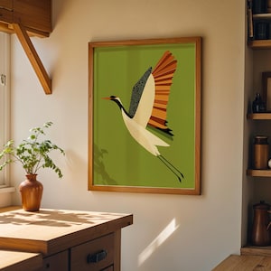 以下が含まれることがあります： 緑色の背景に、空を飛ぶ様式化された鶴を描いた額入りのアートプリント。鶴は黒、白、オレンジ色のディテールが特徴です。このアートワークは木製のフレームに収められており、部屋に自然な暖かさを加えています。
