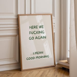 以下が含まれることがあります： 「HERE WE FUCKING GO AGAIN」と「I MEAN GOOD MORNING」の文字が緑色で書かれたフレーム入りのプリント。プリントは薄いベージュ色の背景と木製のフレームで構成されています。アートワークは白い壁に立てかけられています。