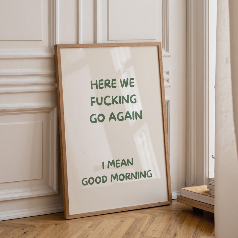 以下が含まれることがあります： 「HERE WE FUCKING GO AGAIN」と「I MEAN GOOD MORNING」の文字が緑色で書かれたフレーム入りのプリント。プリントは薄いベージュ色の背景と木製のフレームで構成されています。アートワークは白い壁に立てかけられています。