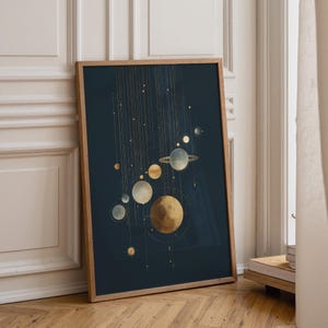 May include: A rectangular framed wall art print featuring a dark navy blue background with a vertical arrangement of golden and pale blue celestial bodies, including planets and rings, connected by thin, delicate vertical golden lines.