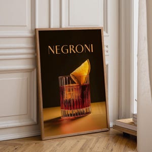 Puede incluir: Un cuadro vertical enmarcado con fondo oscuro y la palabra NEGRONI en letras mayúsculas doradas. Debajo del texto hay un vaso de cóctel rojo con un cubo de hielo grande y una rodaja de naranja, colocado sobre una superficie amarilla.