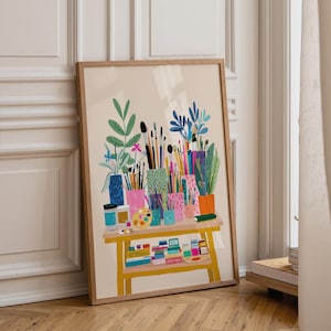May include: A framed art print showing a vibrant illustration of art supplies. The artwork features paintbrushes, paint tubes, and a palette on a yellow table with a lower shelf. The background is a neutral beige.