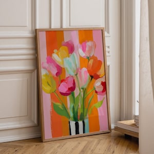 Puede incluir: Una lámina artística enmarcada con un vibrante ramo de tulipanes en tonos rosa, amarillo, naranja y blanco. Las flores están dispuestas en un jarrón de rayas negras y blancas sobre un fondo de rayas verticales naranjas y rosas.