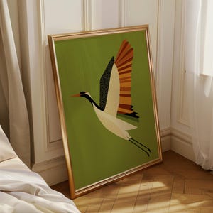 以下が含まれることがあります： 飛行中の様式化された鶴を描いた額入りのアートプリント。鳥は白い体と黒と点々の模様の翼、黒い頭をしています。背景は無地の緑色で、フレームは金色です。