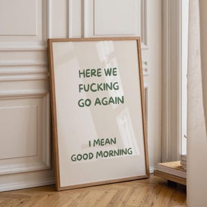May include: A rectangular wall art print in a light wooden frame featuring a cream-coloured background with dark green text that reads: HERE WE FUCKING GO AGAIN I MEAN GOOD MORNING. The print is displayed in a vertical orientation.