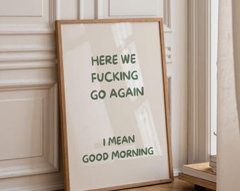 May include: A rectangular wall art print in a light wooden frame featuring a cream-colored background with dark green text that reads: HERE WE FUCKING GO AGAIN I MEAN GOOD MORNING. The print is displayed in a vertical orientation.
