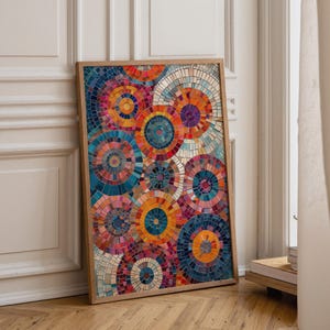 May include: A framed artwork featuring a mosaic design of colorful concentric circles. The circles are composed of small, irregular shapes in shades of blue, orange, red, purple, and white. The artwork is displayed on a wooden floor, leaning against a white wall.