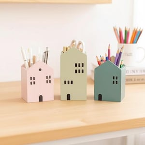 Puede incluir: Tres organizadores de escritorio con forma de casa en colores pastel: rosa, verde y azul. Cada uno tiene una puerta y ventanas recortadas, y contiene bolígrafos, tijeras y lápices de colores. Los organizadores son de madera y están sobre una superficie de madera clara.