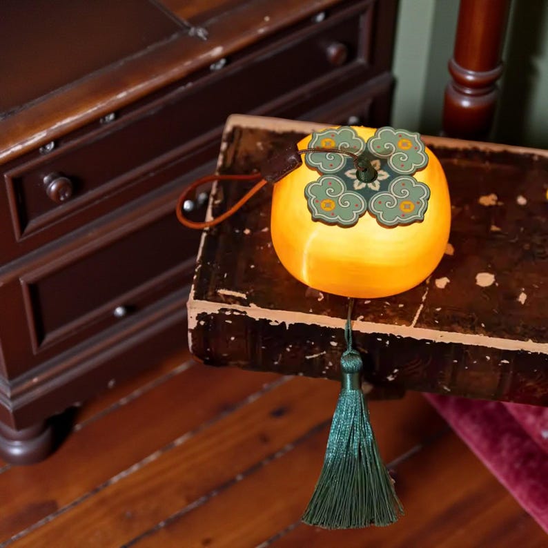May include: A decorative, yellow, spherical object with a green and gold cloud design on top. It has a brown leather strap and a long, green tassel hanging from the bottom. The object is placed on a vintage, dark-colored surface.