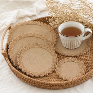 May include: A set of round, tan fabric coasters featuring a decorative scalloped edge made of braided trim. The coasters are arranged in a stack and individually on a woven surface, with one coaster supporting a white ceramic mug filled with tea.