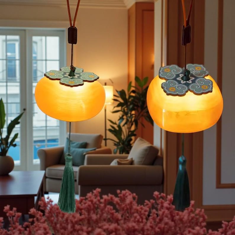 May include: Two hanging pendant lights with a unique design. Each light has a round, orange-yellow shade with a decorative top and a green tassel. The lights are suspended from the ceiling by a brown cord, adding a touch of elegance to the room.