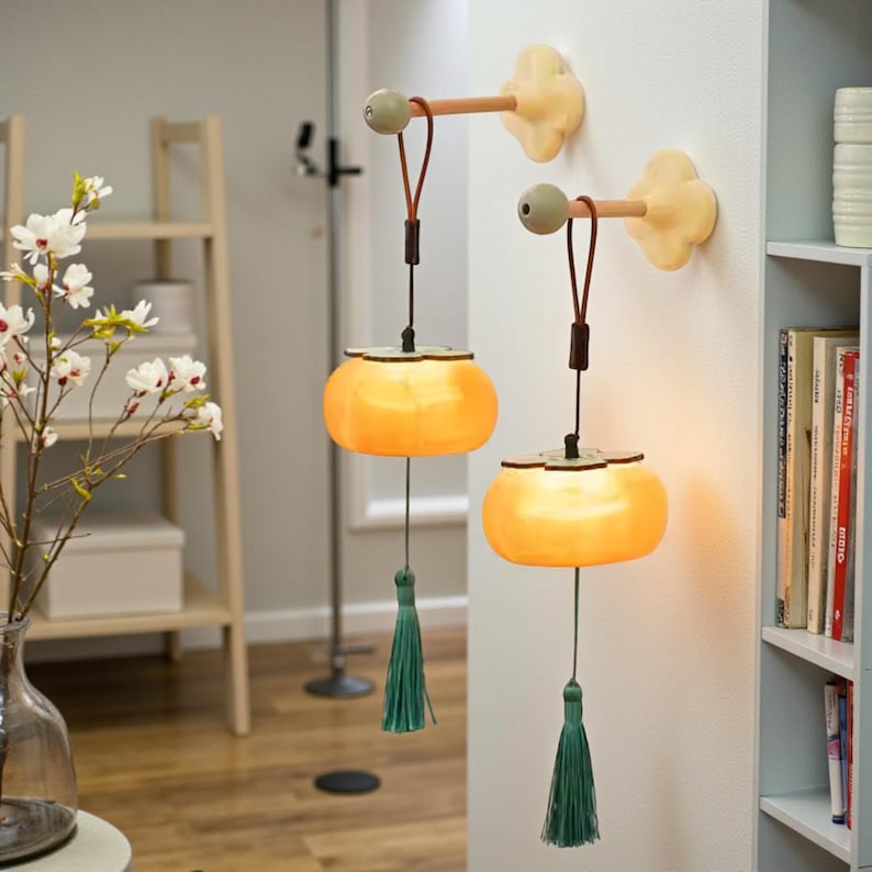 May include: Two wall-mounted, decorative lamps with a warm, orange glow. Each lamp features a round, lantern-like shade, a green tassel, and a wooden arm attached to a cream-colored flower-shaped wall mount. The lamps are hanging on a white wall.