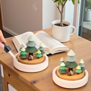 May include: Two decorative forest-themed humidifiers with miniature trees and acorns. The humidifiers are light pink with the words "Breathe Nature" on the base. A hand is using a dropper to add water to one of the humidifiers. A book and a mug are in the background.