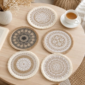 Puede incluir: Cinco posavasos redondos con bordes deshilachados, cada uno con un patrón único de estilo mandala en blanco o negro impreso sobre una superficie de tela color canela. El conjunto incluye varios diseños geométricos y florales complejos.