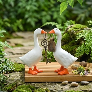 May include: Two white goose figurines with orange beaks and feet, facing each other. They hold a decorative key and a leather keychain. The geese stand on a wooden tray, with a natural background of greenery and small stones.