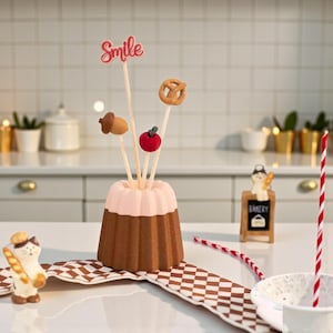May include: A decorative centerpiece with a brown and pink cake-shaped holder and food-themed toppers, including an acorn, pretzel, and red apple. The topper sticks are arranged in a cake-shaped holder. The word "Smile" is written on a topper. A cat figurine with a baguette is on the left.