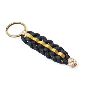 May include: A handmade keychain featuring a braided black and gold design with a brass key ring. The keychain has a tan accent at the end and is a stylish accessory.
