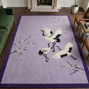 Puede incluir: Una alfombra rectangular lavanda con un borde morado oscuro. La alfombra presenta un diseño de grullas blancas y negras y ramas florecientes. La alfombra está colocada sobre un suelo de madera.