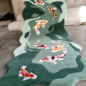 May include: A green and light green rug with a koi fish design. The rug has an irregular shape and features several koi fish in shades of orange, red, black, and white. The rug is made of a soft, plush material.