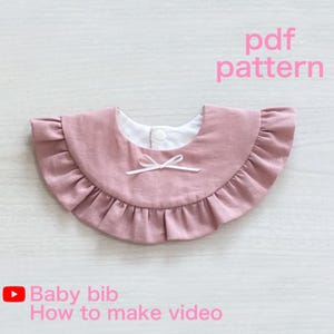 Puede incluir: Babero rosa empolvado con borde con volantes y lazo blanco. El babero tiene interior blanco y cierre de botón. El texto en la imagen dice "pdf pattern" y "Baby bib How to make video".
