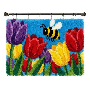 May include: A colorful, handmade wall hanging featuring a vibrant garden scene. The design includes red, yellow, and purple tulips, green leaves, and a cartoon bee against a blue background. The piece is attached to a decorative rod.