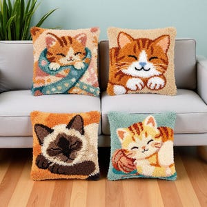 以下が含まれることがあります： パンチニードルで描かれた猫のテクスチャデザインの正方形クッション4点。左上は青いスカーフを巻いた茶トラ猫、右上は茶トラ猫、左下はシャム猫、右下は毛糸玉と一緒の茶トラ猫がそれぞれデザインされています。