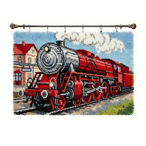 May include: A latch hook wall hanging featuring a red steam locomotive on train tracks. The train emits white smoke against a light blue sky. A building with a red roof is visible in the background. The piece is suspended from a dark metal rod.