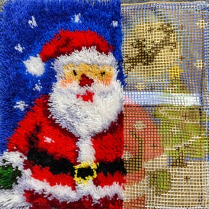 Puede incluir: Un proyecto de manualidades con t&eacute;cnica de gancho que muestra una figura de Santa Claus parcialmente terminada con traje rojo, barba blanca y fondo azul con copos de nieve, junto a una secci&oacute;n inacabada con el lienzo de rejilla.