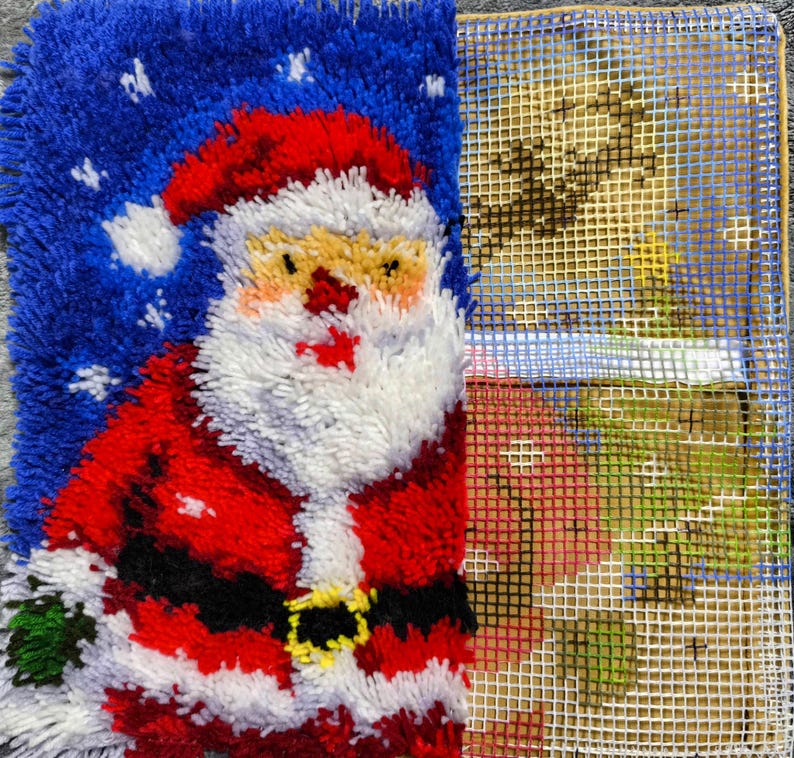 Puede incluir: Un proyecto de manualidades con t&eacute;cnica de gancho que muestra una figura de Santa Claus parcialmente terminada con traje rojo, barba blanca y fondo azul con copos de nieve, junto a una secci&oacute;n inacabada con el lienzo de rejilla.