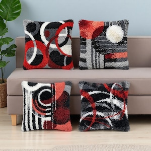 May include: Four square throw pillows with abstract designs in red, black, white, and gray. The pillows have a textured, shaggy appearance and are arranged on a beige sofa. The designs include circles, lines, and geometric shapes.