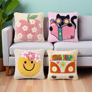 May include: Four decorative throw pillows with embroidered designs. One features a pink apple with white flowers, another a black cat reading a book, a third a yellow smiley face with a flower crown, and the last a colorful van.