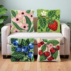 Peut inclure: Quatre coussins carrés avec des motifs en punch needle texturé. L'un présente des fleurs roses, un autre une fraise rouge avec des feuilles vertes, un troisième des myrtilles bleues et le dernier des cerises rouges sur fond crème.
