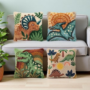 May include: Four decorative throw pillows with dinosaur designs. The pillows feature various dinosaurs in shades of green, brown, and blue, set against backgrounds of beige, orange, and green. The pillows are arranged on a gray sofa.