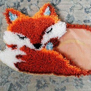 May include: A handmade fox-shaped rug with orange, white, and blue accents. The rug features a detailed design with a textured, tufted appearance. The fox is depicted in a curled-up position, with closed eyes, and measures approximately 18 inches in length.