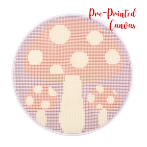 May include: A round, pre-printed canvas featuring a mushroom design. The mushroom has a red cap with white spots and a cream-colored stem. The background is a grid of purple and red threads, with the text "Pre-Printed Canvas" in red.