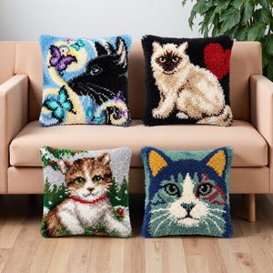 Könnte beinhalten: Vier quadratische Punch-Needle-Kissen mit Katzenmotiven. Eines zeigt eine schwarze Katze mit blauen Schmetterlingen, eines eine beige Katze mit rotem Herz, eines ein Kätzchen im Wald und eines ein blau-weißes Katzengesicht mit gelben Ohren.