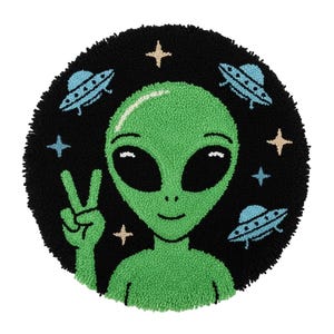 Pode incluir: Um tapete redondo preto com um alienígena verde fazendo o sinal de paz. OVNIs azuis e estrelas circundam o alienígena. O tapete tem uma aparência texturizada e tufada.