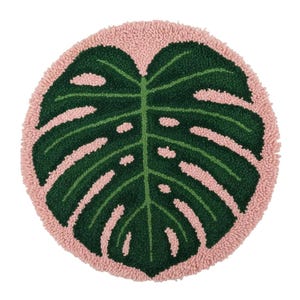 Könnte beinhalten: Runder, getufteter Teppich mit einem großen, grünen Monstera-Blatt-Design und einem rosa Rand. Das Blatt hat mehrere ovale Aussparungen. Der Hintergrund ist weiß.