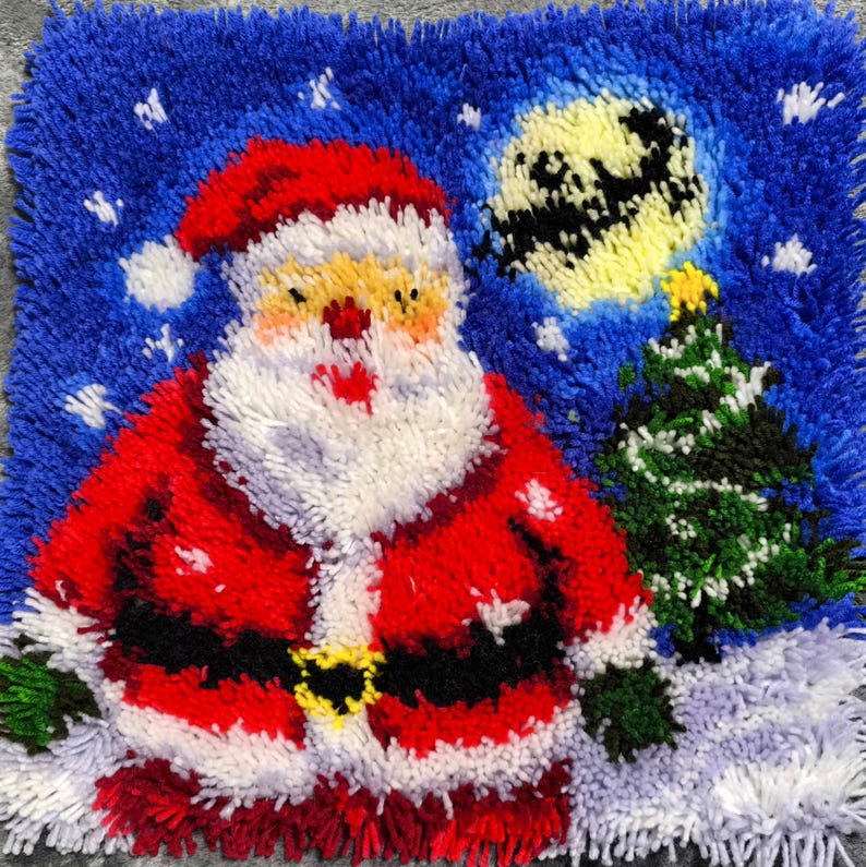 Puede incluir: Una alfombra de ganchillo cuadrada que presenta una figura central de Santa Claus con traje rojo y barba blanca, sobre un cielo nocturno azul con una luna amarilla y un &aacute;rbol de Navidad verde. El dise&ntilde;o incluye detalles de nieve blanca.