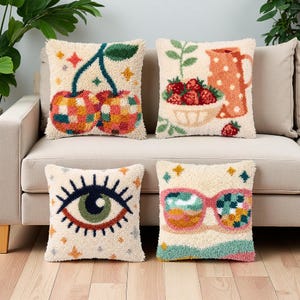 Peut inclure: Quatre coussins carrés en tissu touffeté couleur crème. En haut à gauche, un motif de cerises à carreaux, en haut à droite, un bol de fraises avec une carafe, en bas à gauche, un grand œil, et en bas à droite, une paire de lunettes de soleil.