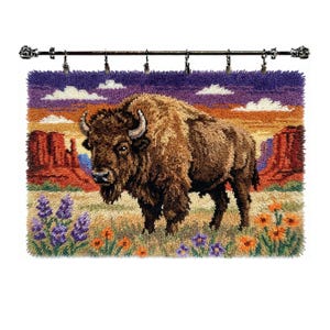 May include: A rectangular latch hook wall hanging featuring a brown bison standing in a desert landscape with red rock formations, purple and orange sunset sky, and foreground patches of purple and orange wildflowers, suspended from a metal rod.