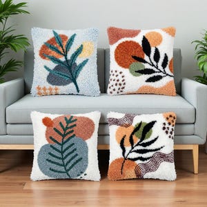 Peut inclure: Quatre coussins carrés avec des motifs botaniques abstraits. Chaque coussin présente un fond blanc avec des motifs colorés et texturés et des motifs de feuilles dans des tons de bleu, orange, vert et marron. Les coussins sont disposés sur un canapé gris.