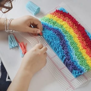 Puede incluir: Un proyecto de manualidades con ganchillo que presenta un vibrante patr&oacute;n de arco&iacute;ris hecho de bucles de hilo multicolor sobre un lienzo de rejilla blanco. El dise&ntilde;o incluye rayas horizontales de hilo azul, morado, amarillo y rojo.