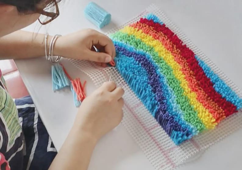Puede incluir: Un proyecto de manualidades con ganchillo que presenta un vibrante patr&oacute;n de arco&iacute;ris hecho de bucles de hilo multicolor sobre un lienzo de rejilla blanco. El dise&ntilde;o incluye rayas horizontales de hilo azul, morado, amarillo y rojo.